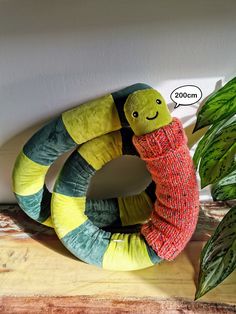 a stuffed snake with a sweater on sitting next to a potted plant and an empty thought bubble