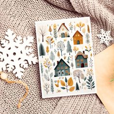 a card with houses surrounded by snowflakes and evergreens on a sweater next to a christmas ornament