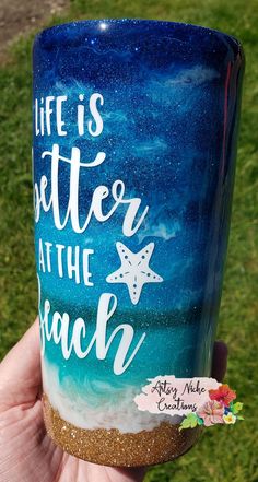 a hand holding up a blue and white coffee cup with the words life is better at the beach painted on it