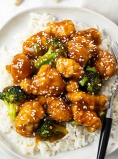 a white plate topped with chicken and broccoli covered in sesame seeds next to rice