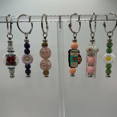 several different colored beads hanging from hooks on a white wall with flowers and charms attached to them