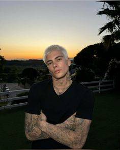 a man with white hair and tattoos standing in front of a fenced area at sunset