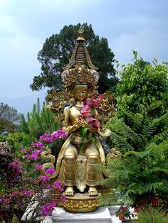 a golden buddha statue sitting in the middle of a garden with purple flowers around it