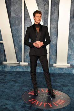 a man in a tuxedo standing on a blue carpet with his hands in his pockets