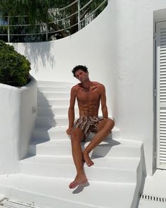 a shirtless man sitting on the steps in front of a white wall and tree