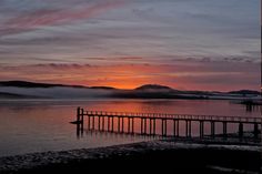 the sun is setting over some water with mountains in the distance and fog on the ground