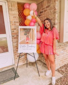 a woman standing in front of a sign with balloons on it and holding up her hand