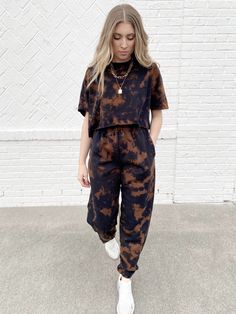 a woman standing in front of a white brick wall wearing a black and brown tie dye jumpsuit