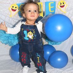 a baby standing in front of blue balloons and smiling at the camera while wearing overalls