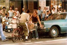 a woman riding on the back of a bike next to a car with people standing around