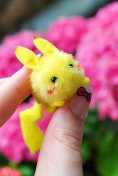 a small yellow stuffed animal sitting on top of someone's finger with pink flowers in the background