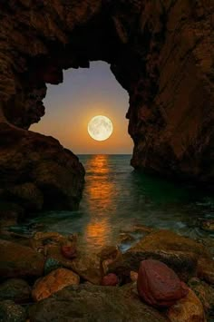 the moon is setting over the ocean with rocks in front of it and an arch leading into