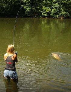 a woman standing in the water while holding a fishing rod with a large fish behind her