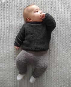 a baby laying on top of a bed wearing a sweater and pants with his hand in his mouth