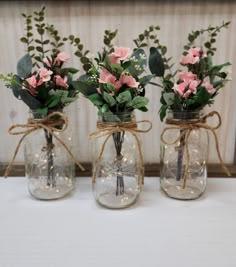 three mason jar vases with white flowers and greenery tied around the top, sitting on a table