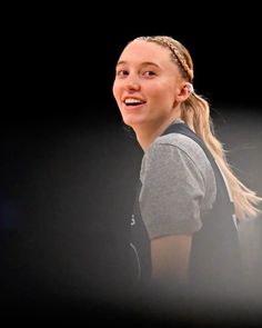 a woman with braids smiles while standing in front of a black background and wearing a gray shirt