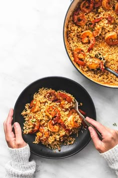 two hands holding a spoon over a plate of shrimp and rice with another pan in the background