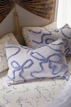 two decorative pillows on a bed with white sheets and blue trimmings, in front of a fan