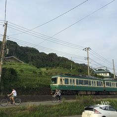 two bicyclists ride past a green train on the tracks in front of a car