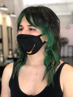 A girl stands in front of an industrial salon background. She has dyed her hair black with green highlighted panels. She is wearing a black tank top and a black face mask. Green Underside Hair, Green Hair Looks, Green Underlayer Hair, Green Tip Hair, Black Hair Green Streaks, Dark Green Money Piece Hair, Under Hair Dye Green, Green Bayalage, Dark Green Hair Streaks