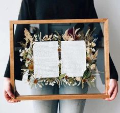 a person holding up a wooden frame with flowers and paper attached to the sides, in front of a white wall