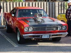 two men standing next to a red and black muscle car