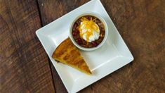 a white plate topped with a piece of cake next to a bowl of chili and whipped cream