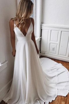 a woman standing in front of a window wearing a white wedding dress with spaghetti straps