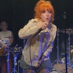 a woman with red hair standing in front of a microphone and drum set on stage