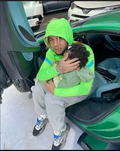 a man in a green hoodie is holding a small dog while sitting on the back of a car