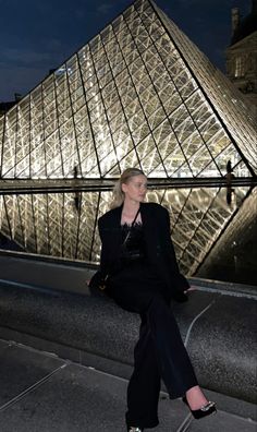 a woman sitting on the edge of a walkway next to a glass pyramid at night