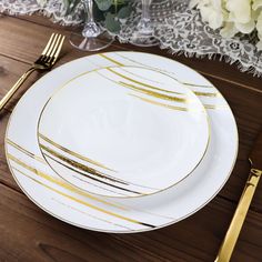 a white plate with gold lines on it next to two forks and a glass vase filled with flowers