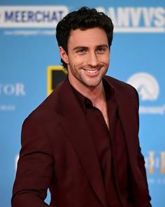 a smiling man in a maroon suit and black shirt smiles at the camera with his hands on his hips