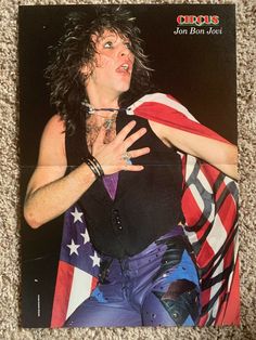 a man with long hair sitting in front of an american flag and holding his arms out