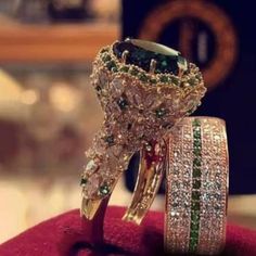 two gold rings with green and white diamonds on top of red velvet covered cushiones