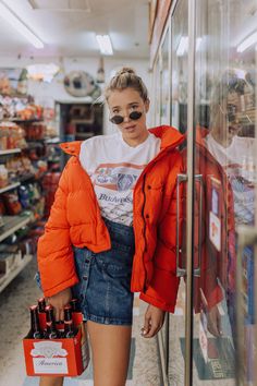 a woman in an orange puffer jacket and black sunglasses is holding a beer bottle