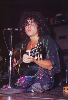 a man sitting on the floor playing an acoustic guitar and singing into a microphone while holding a guitar in his right hand