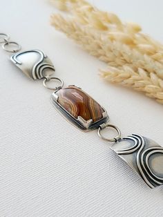 Reminiscent of steep desert canyon walls, slowly cut away by meandering waterways, this bracelet is purely inspired by the layered bands of color winding through the Laguna Lace Agate. All the hues of the desert can be found in this unique stone: red clay, faded green, sandy browns, and sunset purples. The stone is held in place with silver at all four corners so you can see the colors moving through the agate, and a window cut in the back reveals the continuous maze of colorful patterns on the Artisan Sterling Silver Bracelet With Natural Stones, Handmade Jasper Bracelet, Earthy Agate Bracelet Jewelry, Brown Agate Bracelet, Bohemian Jasper Bracelet, Artisan Agate Jewelry With Polished Finish, Silver Artisan Agate Bracelets, Artisan Silver Agate Bracelets, Jasper Natural Stone Bracelet