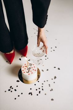 a woman in black pants and red high heels lighting a candle on a white cake