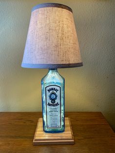 This Bombay Sapphire lamp looks great with our standard tan shade and our new kraft laced shade.  We also have a gray shade as an option and all shades come with a bulb.  Our lamps are high quality hand crafted liquor bottle table lamps made from distinctive bottles repurposed into unique lamps. Made in Tampa Bay, FL, each one is custom made with a Leviton 2-circuit lamp socket fitting a standard size bulb, with LED mini lights inside the bottle, an 8 foot long clear silver lamp cord, and mounted on a stained and beveled oak wood base. The 2-circuit switch allows the user to choose between the top bulb in the socket or the bottom mini lights (nice for a night light) or both at the same time. Repurpose Liquor Bottles, Old Liquor Bottles, Bottle Table Lamps, London Gin, Bombay Sapphire, Silver Lamp, Mini Lights, Bottle Diy, Wine Bottle Diy Crafts