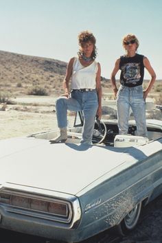 two women standing on top of an old car in the middle of nowhere, with mountains in the background