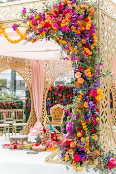 an elaborately decorated table with colorful flowers and cake on the side, in front of a white gazebo