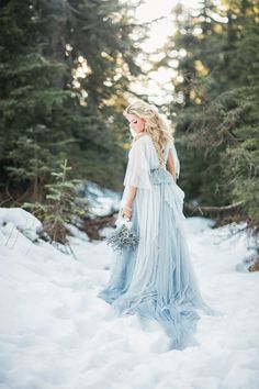 a woman standing in the snow wearing a blue dress and holding a bouquet with trees behind her