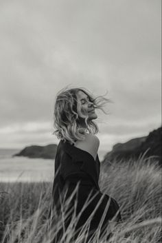 a woman standing in tall grass near the ocean with her hair blowing in the wind