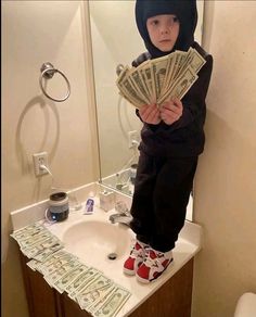 a young boy standing on top of a bathroom counter holding money in front of his face