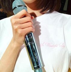 a woman holding a microphone in her right hand and wearing a white shirt with pink writing on it