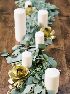 some white candles are lined up on a table with greenery and gold flowers in the middle