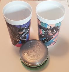 two cups with batman images on them sitting next to an empty cup holder and lid