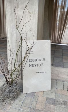 a white sign sitting next to a plant on top of a brick floor in front of a window