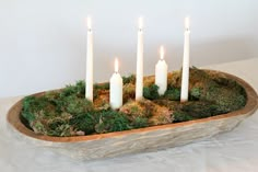 four white candles are placed in a moss covered bowl